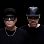 Two men in studio wearing black with black caps and sunglasses