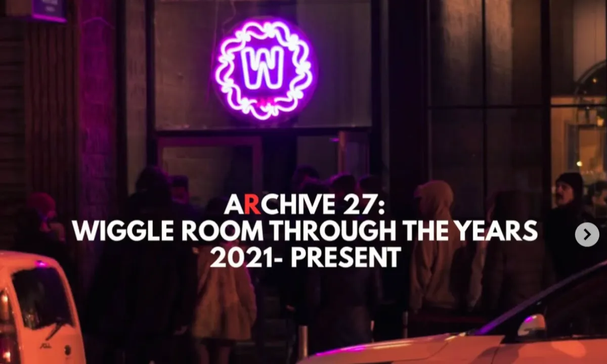 A group of people stands outside a building at night under a glowing purple neon W sign. Text reads: ARCHIVE 27: WIGGLE ROOM THROUGH THE YEARS 2021- PRESENT.