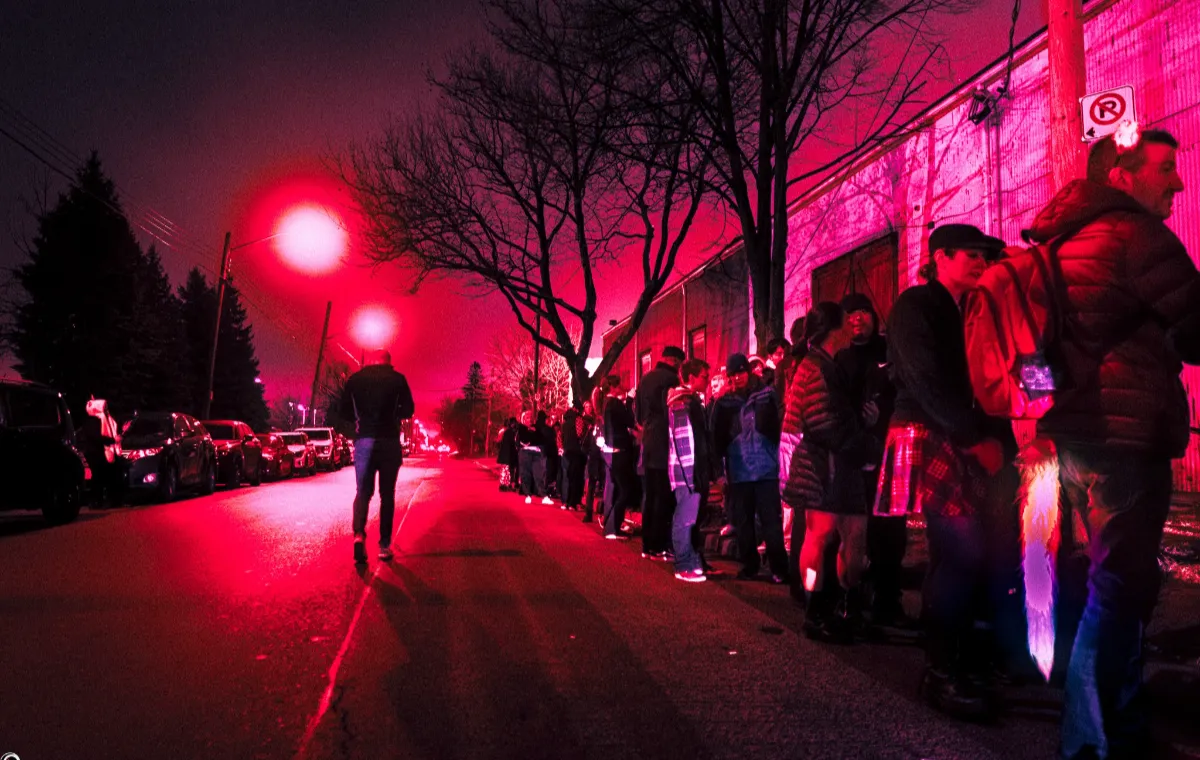 A long line of people waits outside a building at night, illuminated by bright pink and red streetlights. Bare trees and parked cars line the street, and one person walks alone toward the camera.