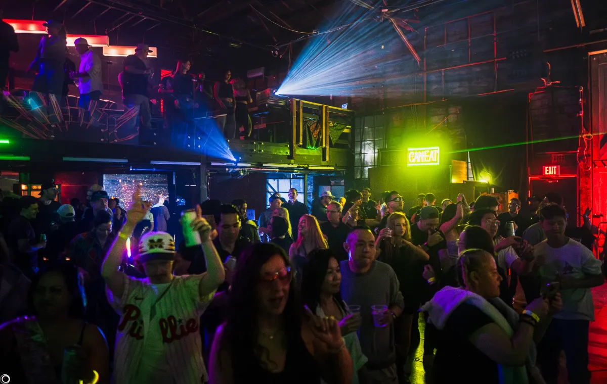 A lively crowd dances and socializes in a dimly lit nightclub with colorful lights and lasers. People hold drinks, a neon CANTEEN sign glows on the wall, and an upper level overlooks the bustling scene below.