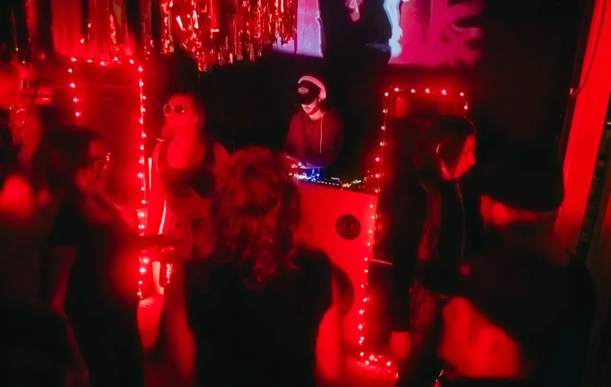 A DJ wearing a cap and sunglasses performs at a booth surrounded by red string lights, while people dance and socialize under red lighting in a dark, decorated room.
