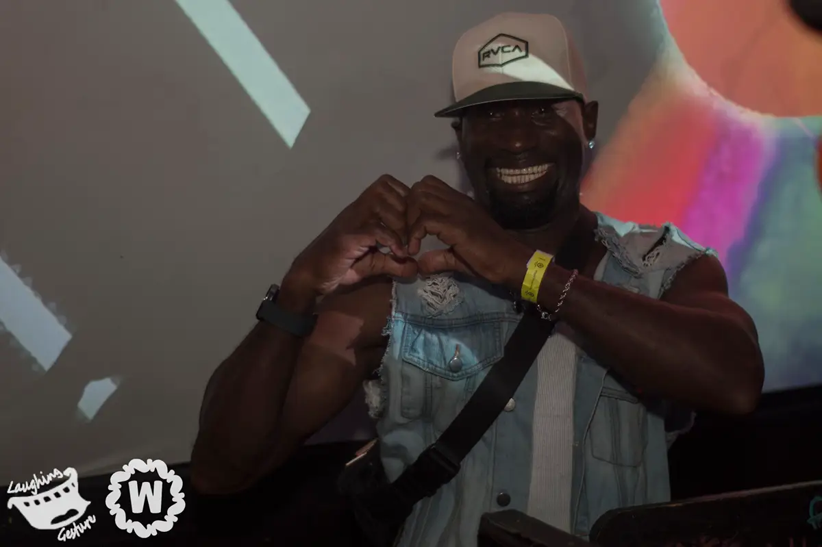 DJ with arms forming a heart shape, wearing a denim vest and RVCA hat.